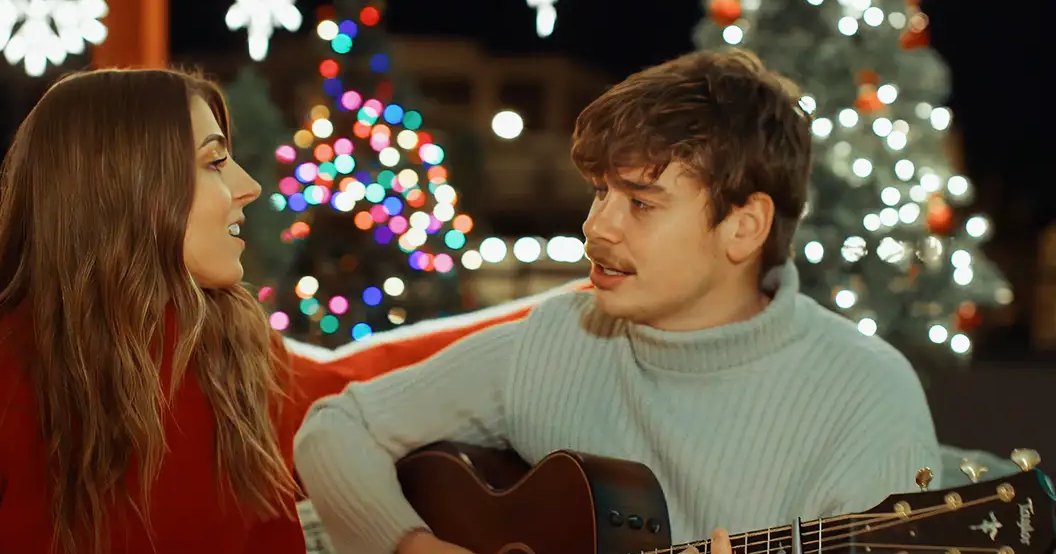 A couple singing in front of decorated Christmas trees at night, with a festive outdoor setting and artificial lighting creating a cozy holiday atmosphere.
