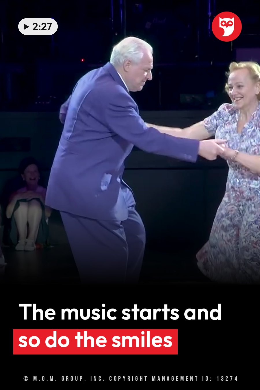 An older man and woman dance together on a stage, smiling and holding hands. A headline runs in white letters across the lower third.