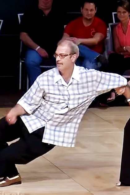 Middle-aged man dancing with an older adult woman in a dance studio or event space, surrounded by chairs and an audience watching.