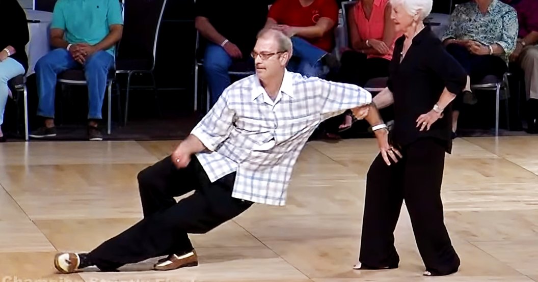 Middle-aged man dancing with an older adult woman in a dance studio or event space, surrounded by chairs and an audience watching.