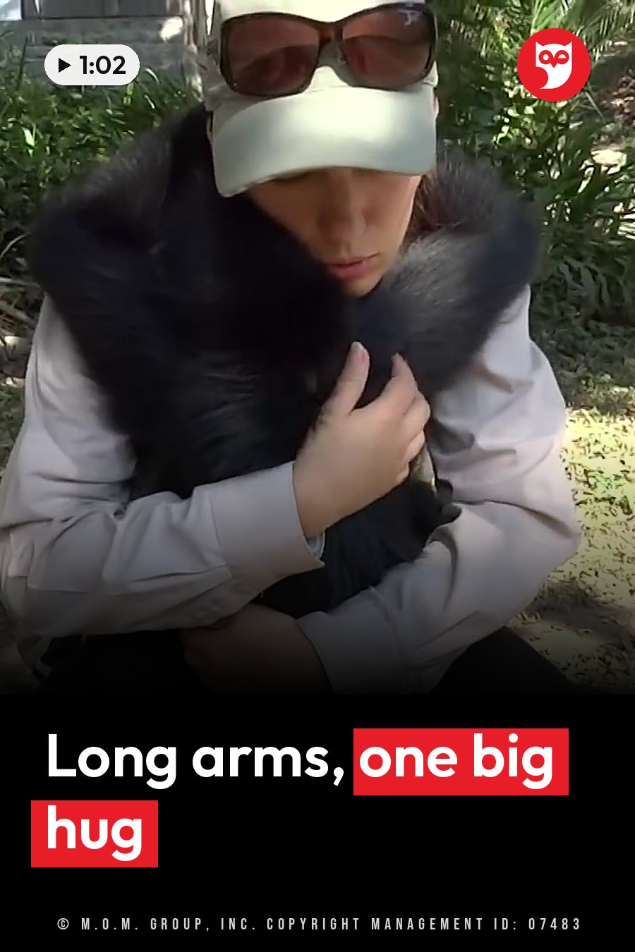 A woman in a cap and sunglasses holds a black-furred monkey close to her chest outdoors. A headline runs in white letters across the lower third.