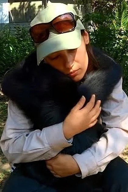 A young adult male wearing a light-colored shirt with a darker vest and dark pants holds a small black dog in a lush garden on a bright sunny day.