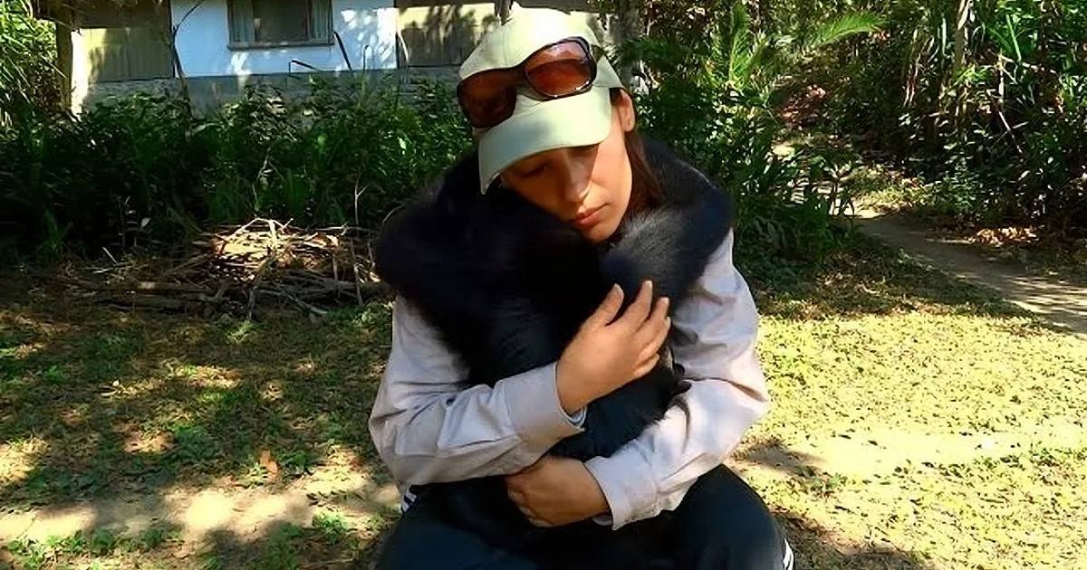 A young adult male wearing a light-colored shirt with a darker vest and dark pants holds a small black dog in a lush garden on a bright sunny day.