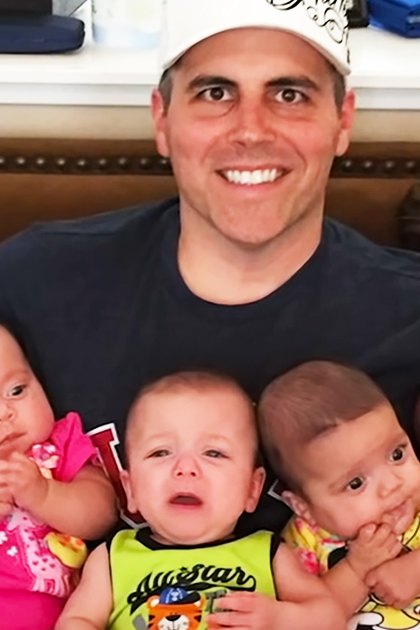 A middle-aged man indoors, surrounded by infants in colorful outfits; brown leather couch with decorative stitching and a white shelf holding various items. Text on clothing: 'Allstar'.