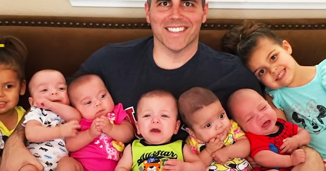 A middle-aged man indoors, surrounded by infants in colorful outfits; brown leather couch with decorative stitching and a white shelf holding various items. Text on clothing: 'Allstar'.