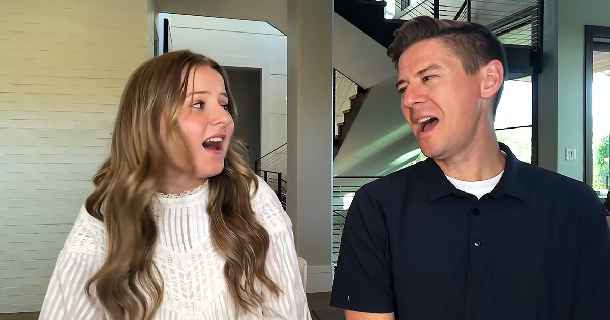 Young adult female in a white lace blouse and young adult male in a dark blue polo shirt indoors, modern home setting with staircase and natural daylight.
