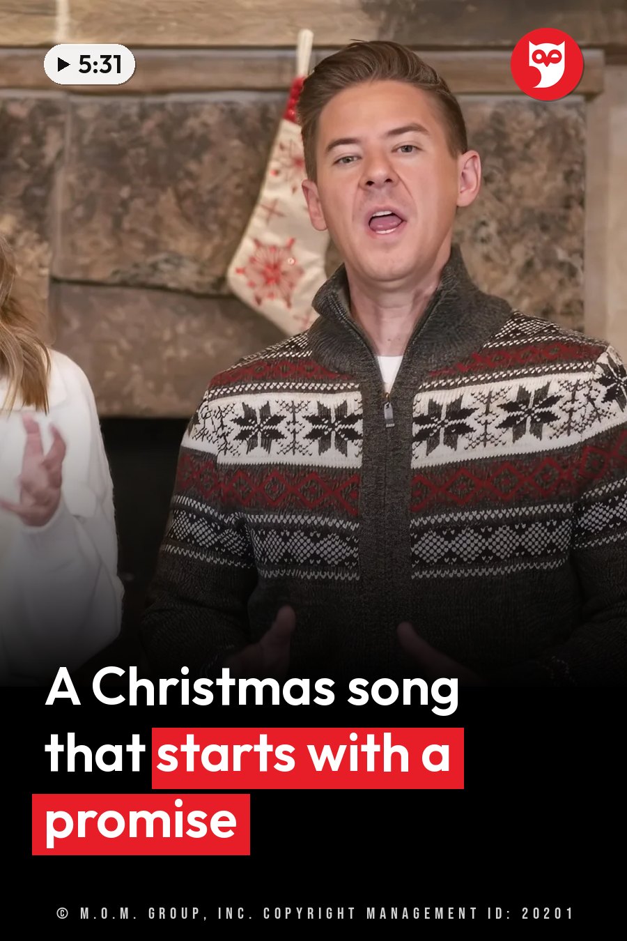 A man in a patterned sweater and a woman beside him sing and gesture near a fireplace with a stocking. A headline runs in white letters across the lower third.