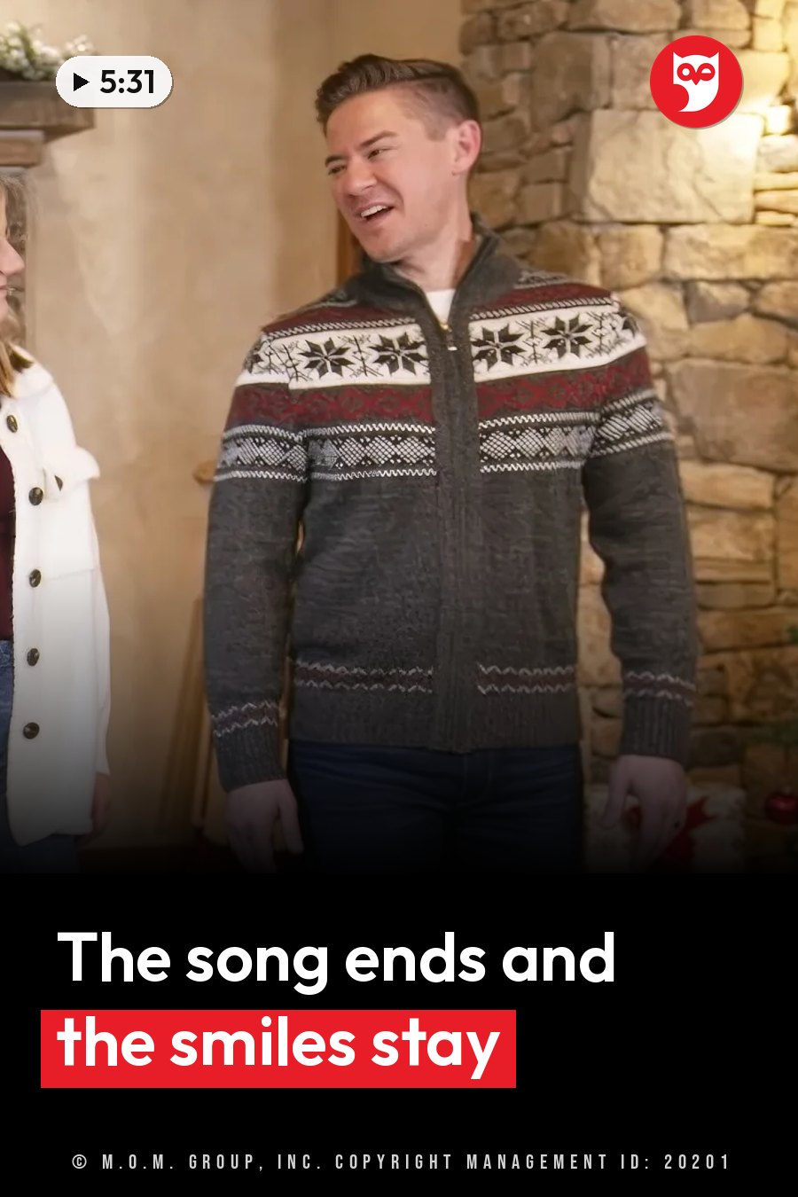 A man in a patterned sweater smiles near a stone wall, with another person partly visible beside him. A headline runs in white letters across the lower third.