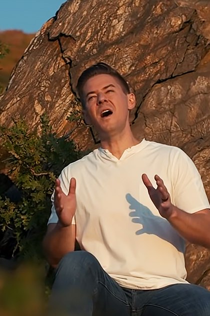 Two young adults, a female in red top and jeans with bracelets, and a male in white polo shirt and jeans, outdoors near rocks and greenery under warm late afternoon or early evening light.