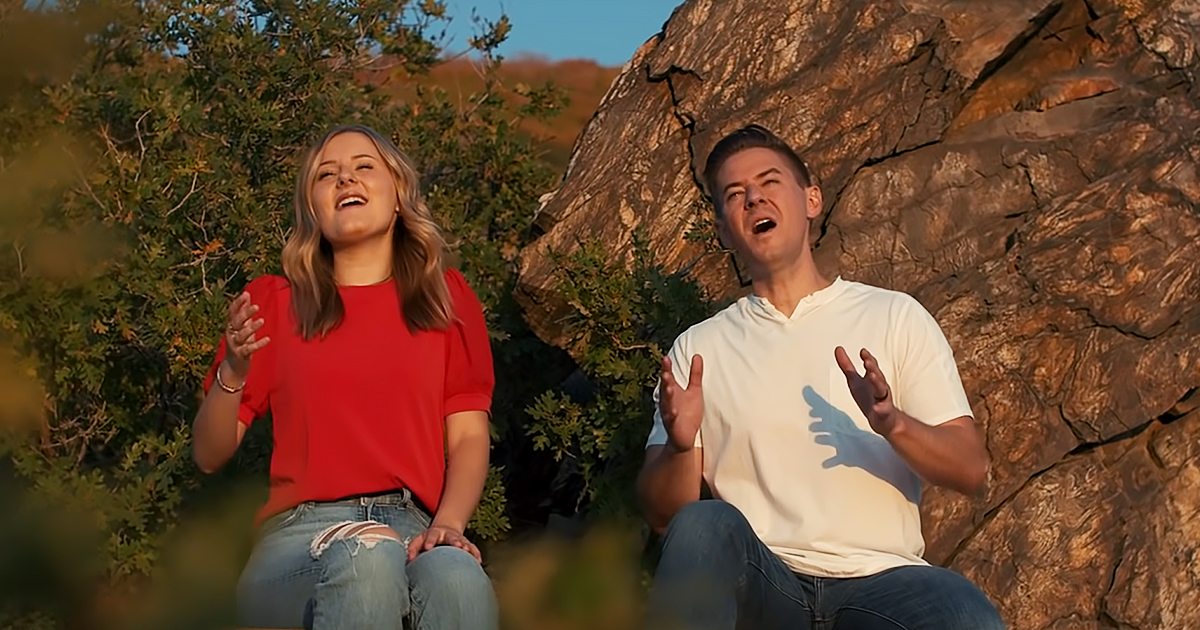 Two young adults, a female in red top and jeans with bracelets, and a male in white polo shirt and jeans, outdoors near rocks and greenery under warm late afternoon or early evening light.