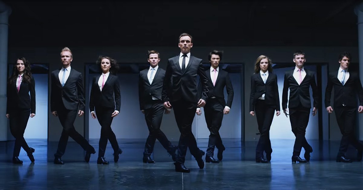 Group of individuals dressed in formal attire, standing indoors with a neutral background and even lighting.