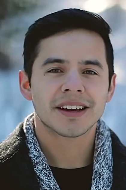 A young adult male stands outdoors in a snowy forest, wearing a dark, textured collar jacket with long sleeves folded up and a blue scarf.