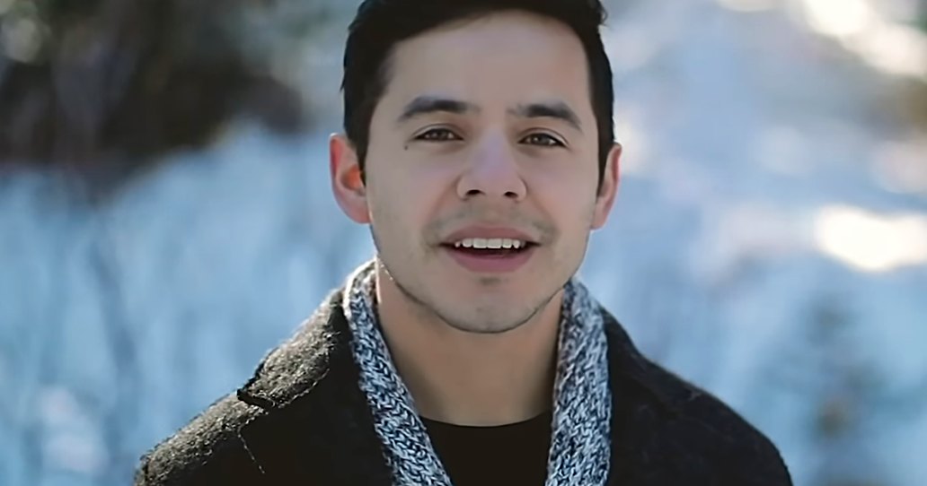 A young adult male stands outdoors in a snowy forest, wearing a dark, textured collar jacket with long sleeves folded up and a blue scarf.