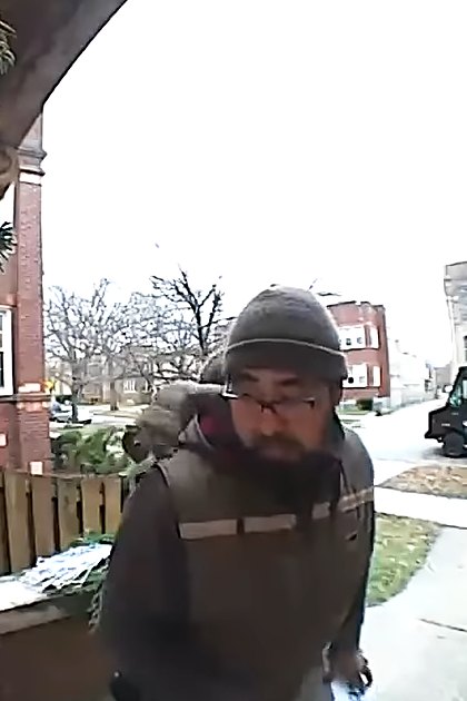A delivery person wearing a dark jacket with light and darker sections, standing at a residential location during winter, with a security camera lens in view.