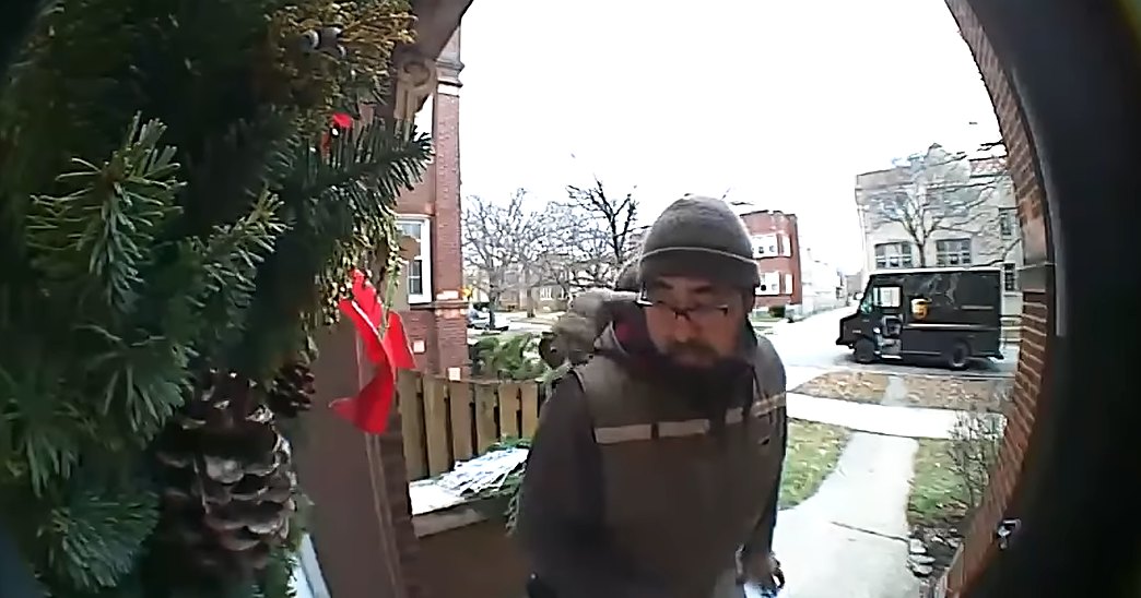 A delivery person wearing a dark jacket with light and darker sections, standing at a residential location during winter, with a security camera lens in view.