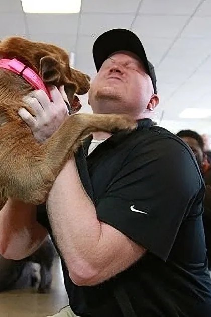 An adult male hugging a golden-brown Labrador Retriever mix in an indoor setting with other people interacting with animals, bright lighting suggesting daytime.