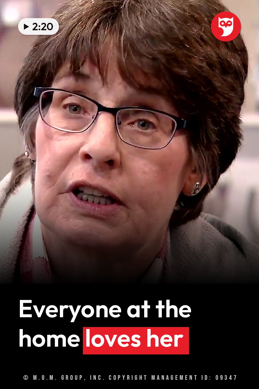 An older woman with glasses, a scarf and a coat speaks. A headline runs in white letters across the lower third.