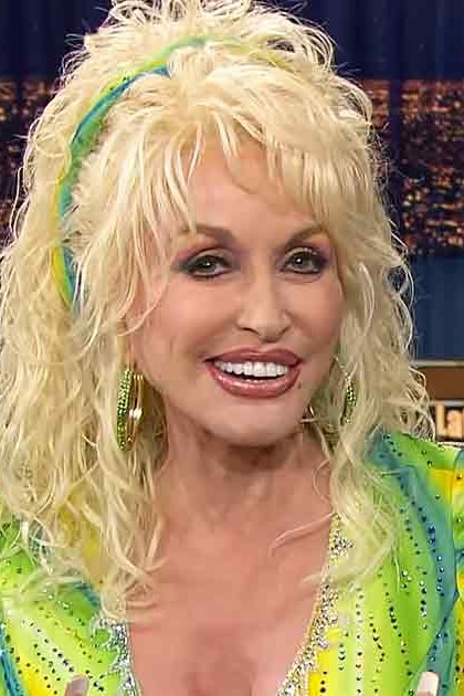A middle-aged woman in a vibrant green and yellow patterned sequined dress on a late-night talk show set with urban night backdrop.