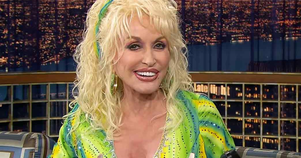 A middle-aged woman in a vibrant green and yellow patterned sequined dress on a late-night talk show set with urban night backdrop.