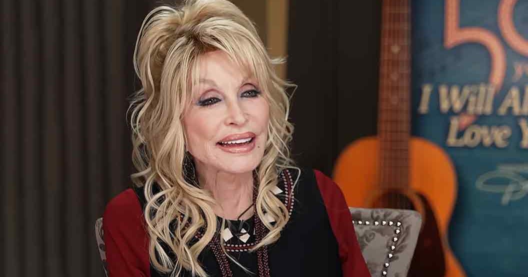 A middle-aged woman in a black top with intricate patterns, paired with a red cardigan or jacket, seated indoors with decorative furniture and a guitar in the background.