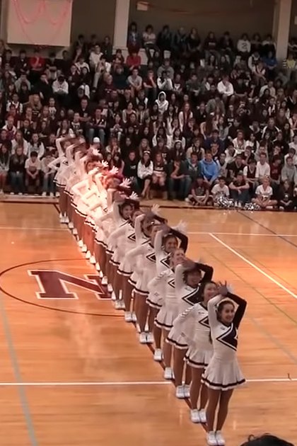 Cheerleaders performing in a gymnasium with an audience watching, indoors during daytime or early evening under indoor lighting.