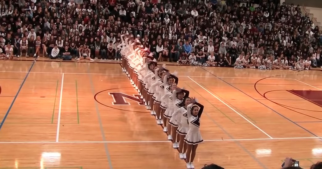 Cheerleaders performing in a gymnasium with an audience watching, indoors during daytime or early evening under indoor lighting.
