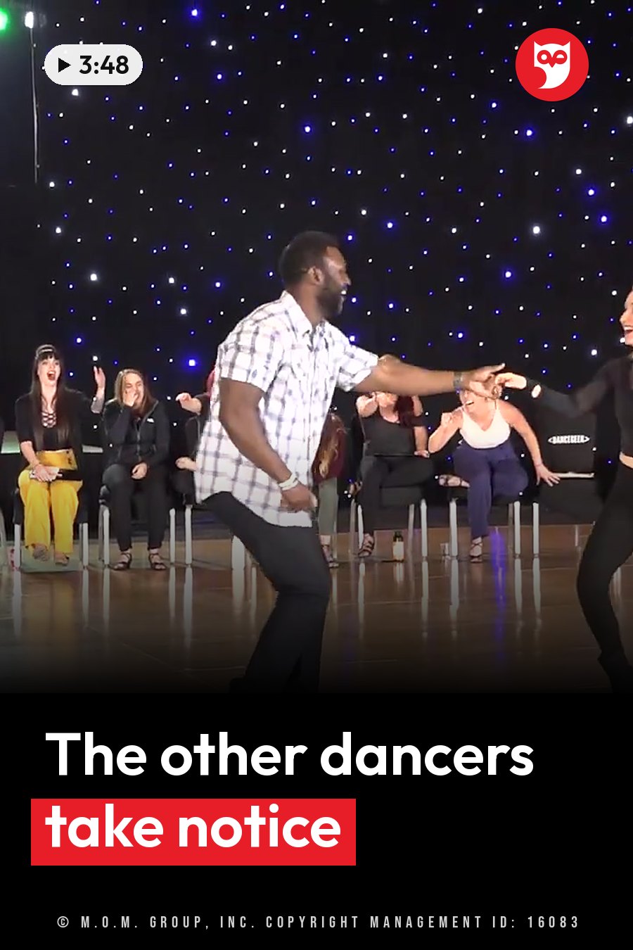 A man dances on a stage with a woman while a seated audience watches and reacts. A headline runs in white letters across the lower third.