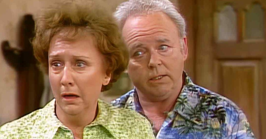 A middle-aged woman in a green patterned shirt stands indoors, accompanied by a middle-aged man wearing a blue palm tree-patterned shirt. The setting appears to be an older home interior with wooden doors and textured walls.