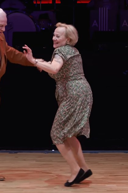 An older adult male and female couple dancing indoors, the man wearing a brown suit with white dress shoes, the woman in a green patterned dress with black slip-on shoes.