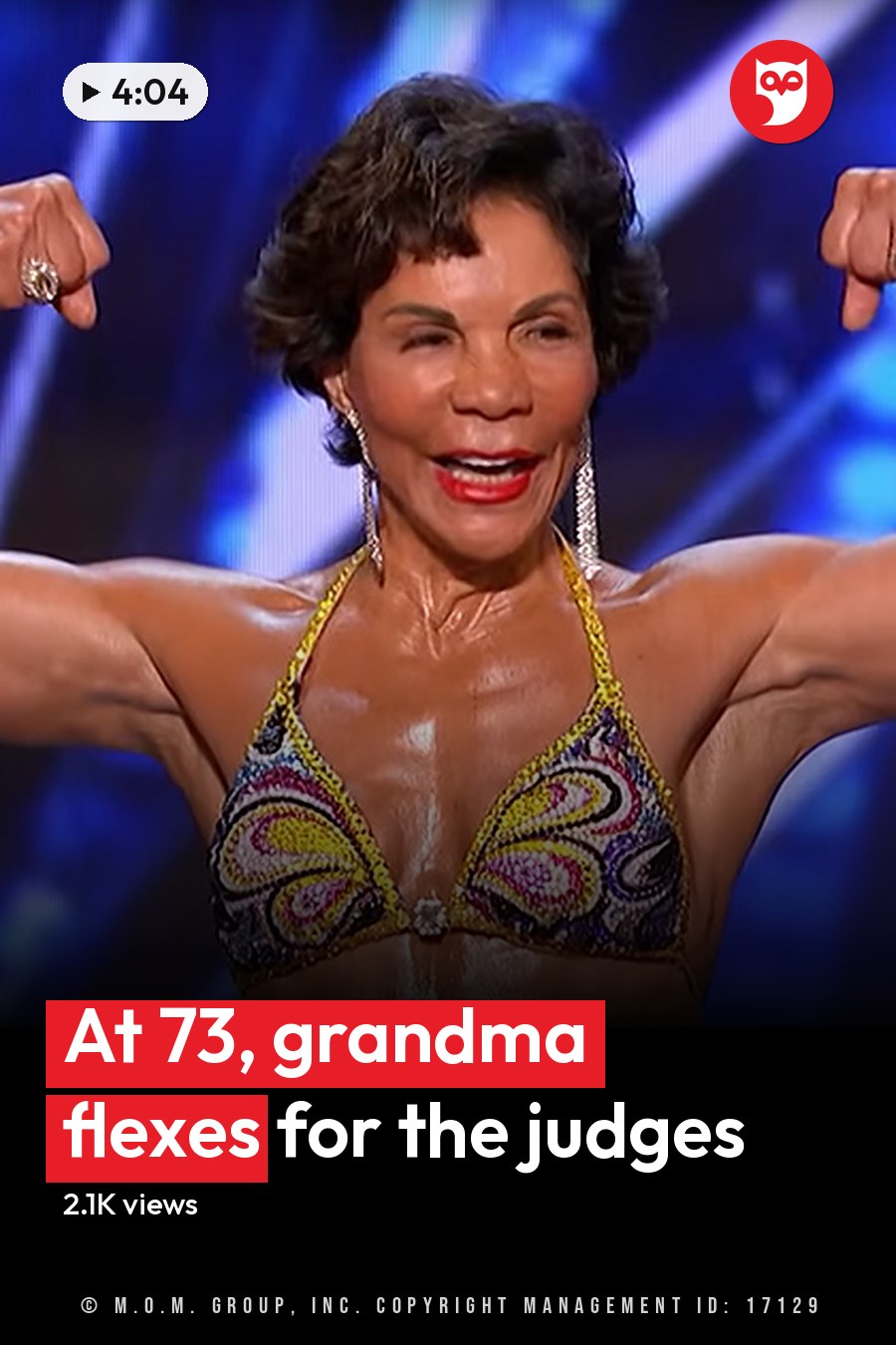 A middle-aged adult female in a form-fitting, bright multicolored bikini with intricate patterns and accessories stands on stage at an indoor fitness competition under geometric blue lighting. The headline runs in white letters across the lower third.