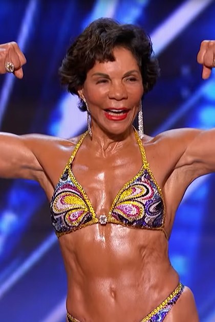 A middle-aged adult female in a form-fitting, bright multicolored bikini with intricate patterns and accessories stands on stage at an indoor fitness competition under geometric blue lighting.