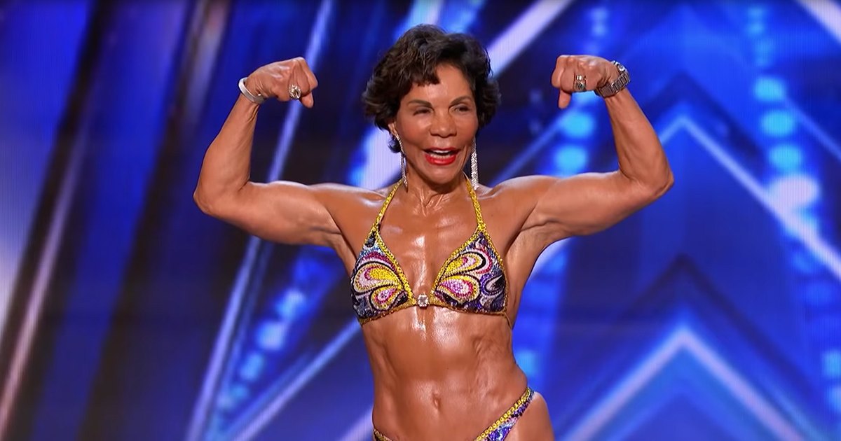 A middle-aged adult female in a form-fitting, bright multicolored bikini with intricate patterns and accessories stands on stage at an indoor fitness competition under geometric blue lighting.