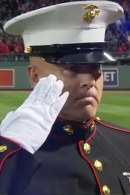 An adult male marine saluting in Fenway Park during a spring or summer baseball game with a large crowd of spectators in the background.