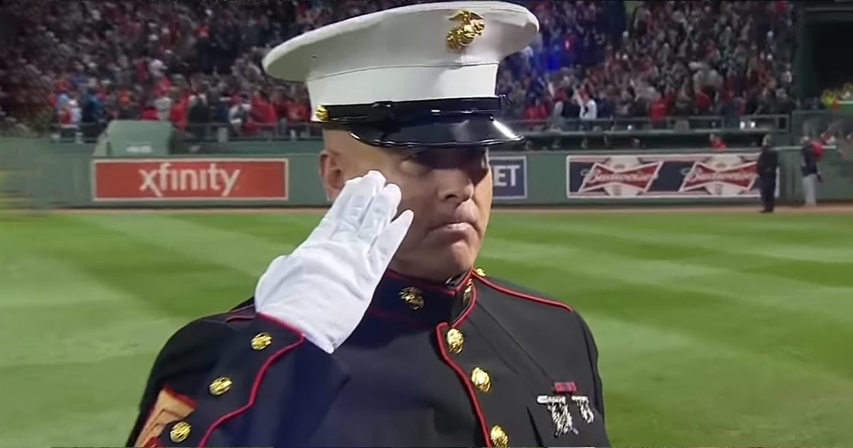 An adult male marine saluting in Fenway Park during a spring or summer baseball game with a large crowd of spectators in the background.