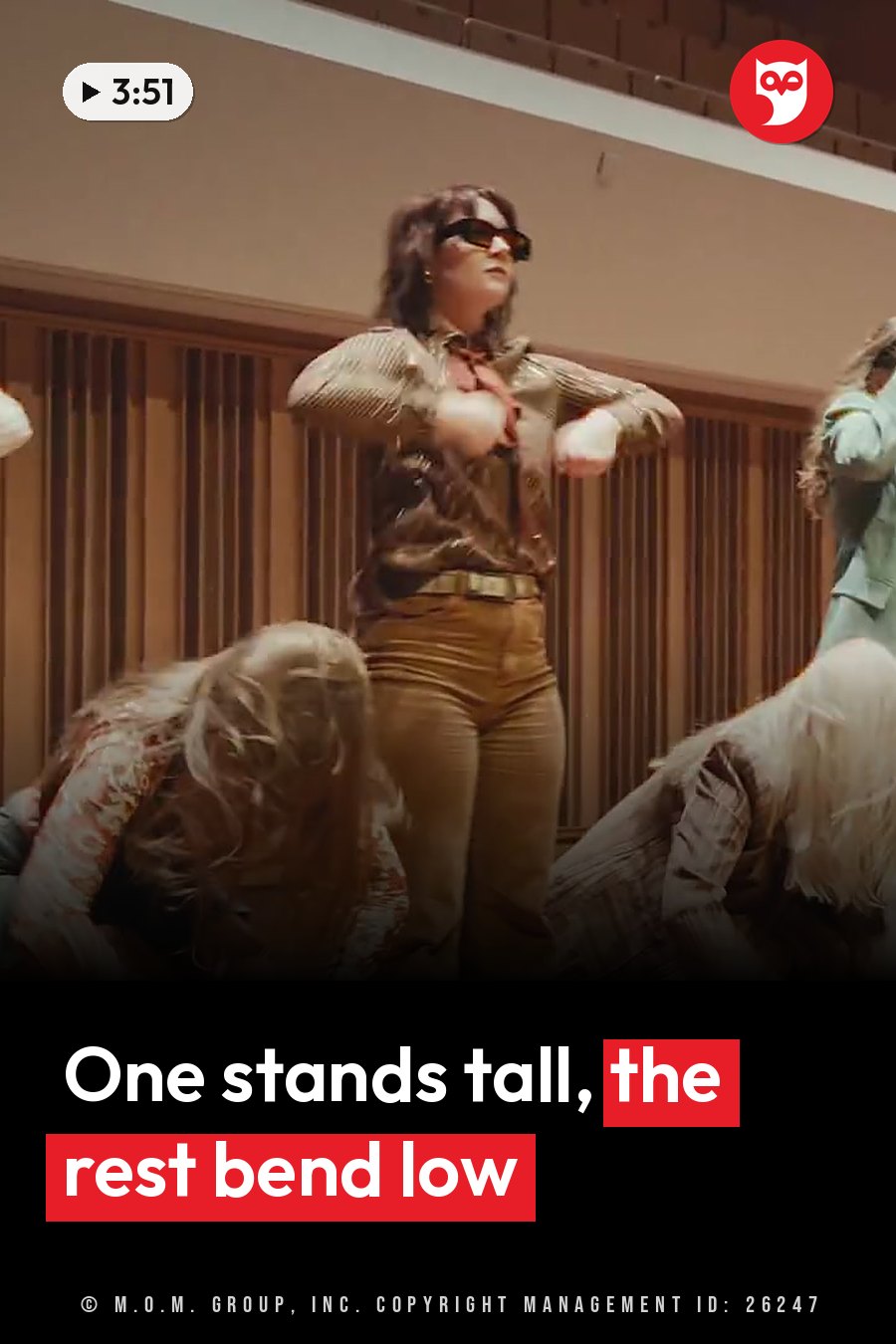 A person in sunglasses and a shirt stands with arms bent while other people bend down nearby, indoors. A headline runs in white letters across the lower third.