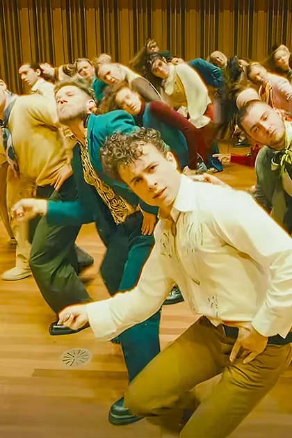 A young adult male stands in an indoor space with wooden flooring, wearing a white long-sleeve shirt with rolled-up sleeves and brown pants, amidst a group of formally dressed individuals.