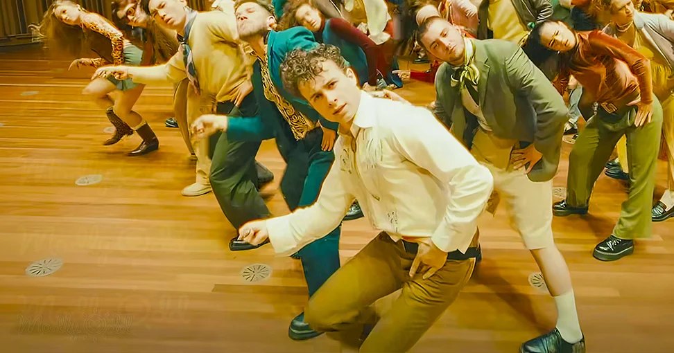 A young adult male stands in an indoor space with wooden flooring, wearing a white long-sleeve shirt with rolled-up sleeves and brown pants, amidst a group of formally dressed individuals.