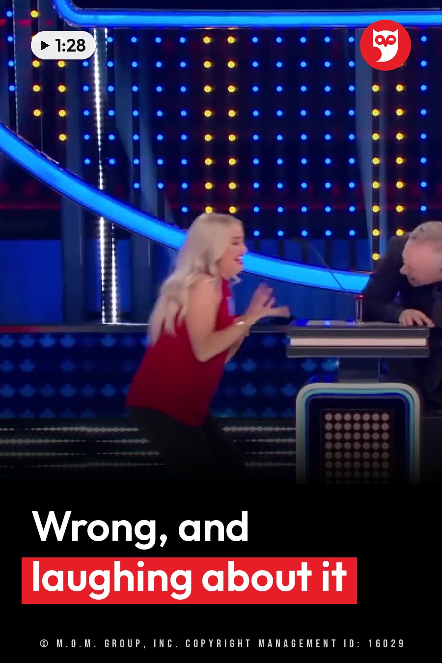 A woman in a red shirt laughs and gestures toward a man seated at a desk on a brightly lit stage. A headline runs in white letters across the lower third.
