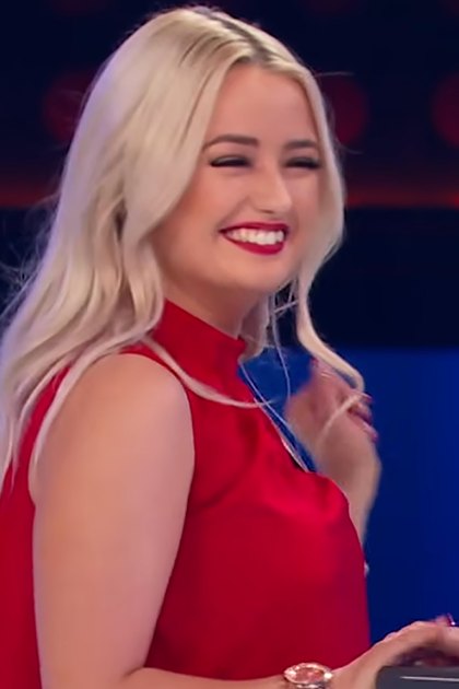 A young adult female in a bright red satin dress stands on an indoor stage with a microphone, suggesting a performance or competition setting.