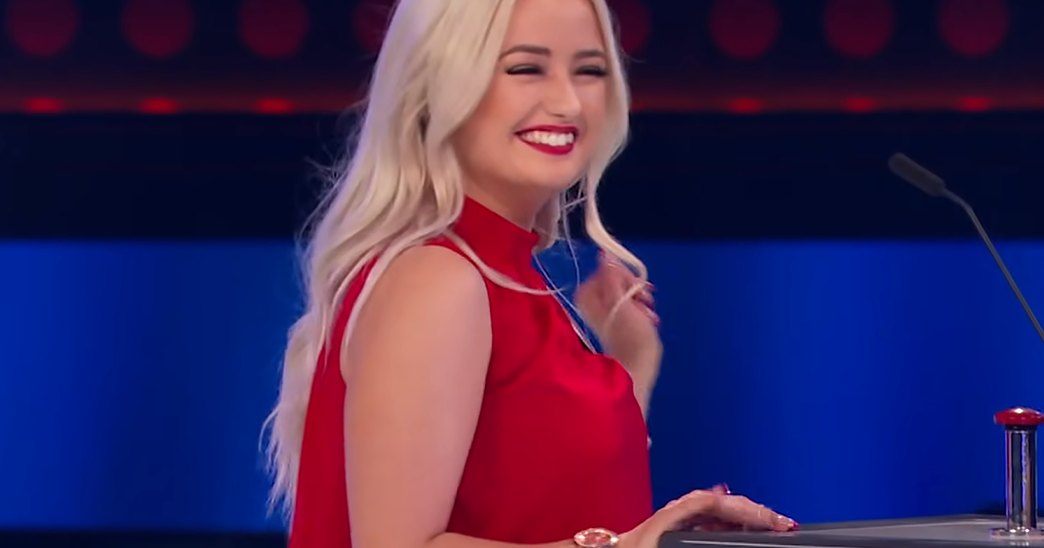 A young adult female in a bright red satin dress stands on an indoor stage with a microphone, suggesting a performance or competition setting.