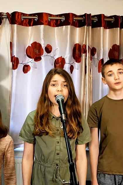 A family sings together in a home setting with curtains with red floral patterns, suggesting spring or summer. The scene is well-lit by natural daylight.
