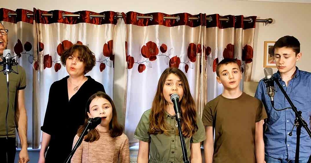 A family sings together in a home setting with curtains with red floral patterns, suggesting spring or summer. The scene is well-lit by natural daylight.