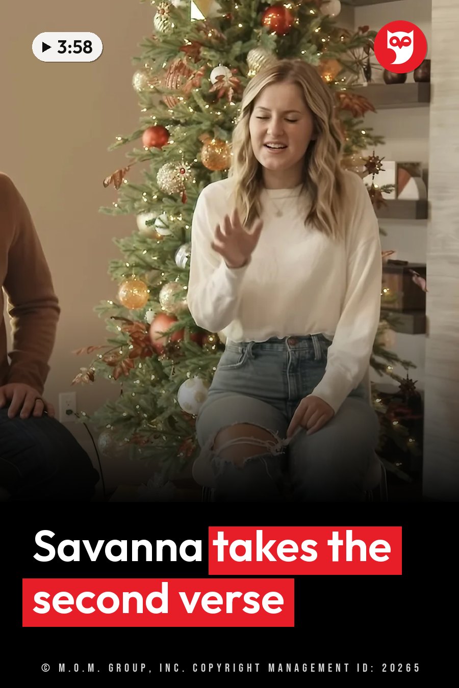 A young woman sits near a decorated Christmas tree, smiling slightly and gesturing with one hand. A headline runs in white letters across the lower third.