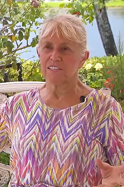 An older adult female in a casual, contemporary zigzag-patterned blouse sits on a white metal bench with a light golden Labrador Retriever mix wearing a collar and tag amidst lush green foliage in bright daylight, suggesting spring or summer.