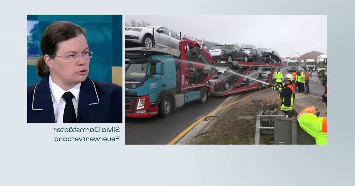 An adult female in a professional interview setting for emergency services, with multiple vehicles including a car transporter truck on an outdoor highway under diffused natural light.