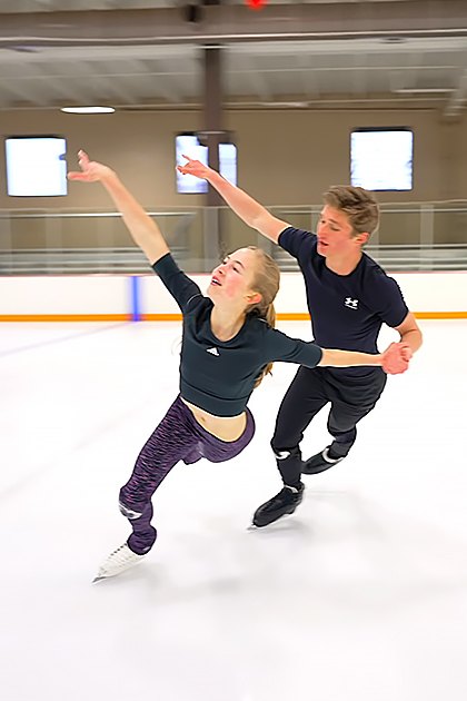 A young couple ice skating indoors at an indoor rink with white flooring and orange boundary lines, natural light from windows indicating daytime.