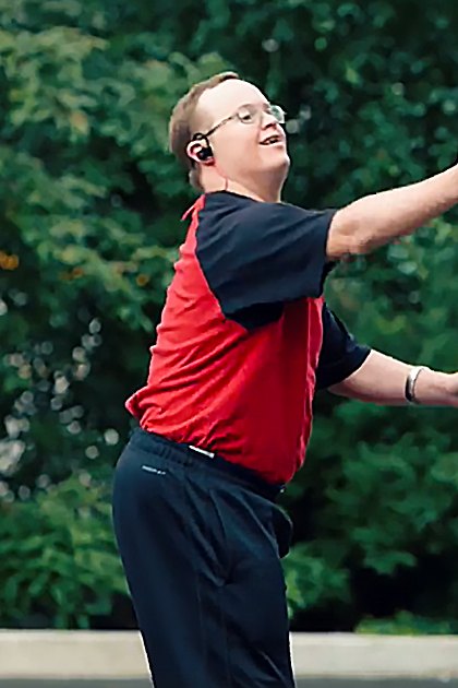 A young adult male wearing a red and black short-sleeved shirt with dark athletic or casual pants, glasses, a watch, and an earpiece in a park-like outdoor setting with green trees and soft lighting.