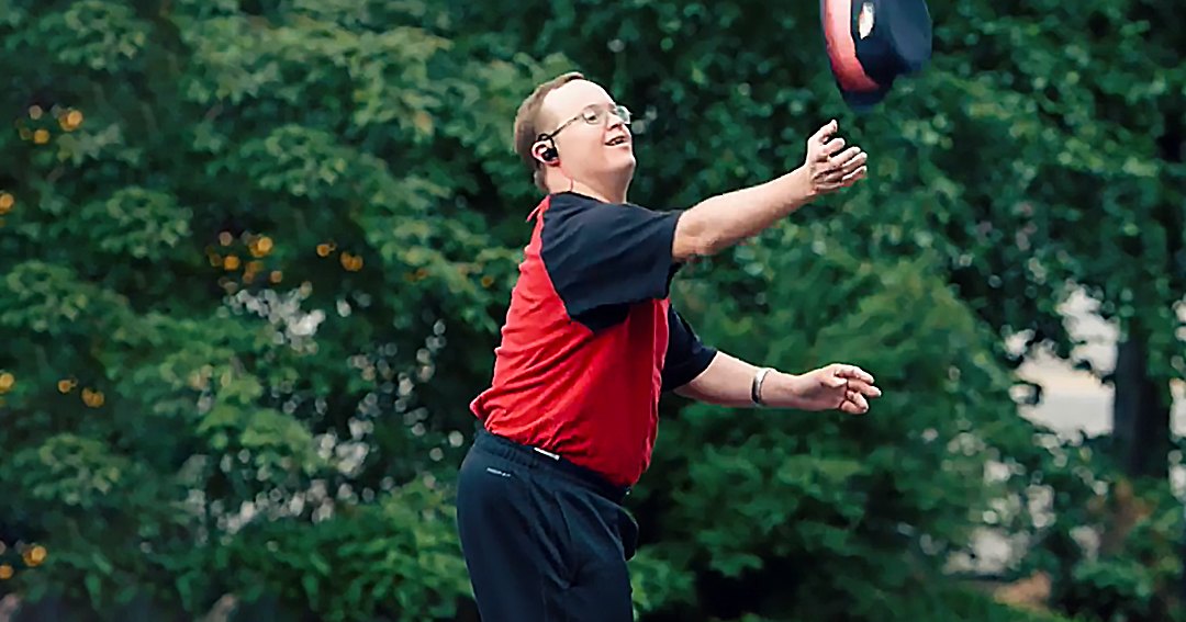 A young adult male wearing a red and black short-sleeved shirt with dark athletic or casual pants, glasses, a watch, and an earpiece in a park-like outdoor setting with green trees and soft lighting.