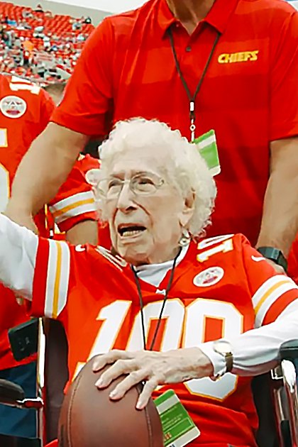 An older adult woman in a stadium wearing a red Kansas City Chiefs jersey with the number 100, surrounded by other individuals also dressed for a football game.