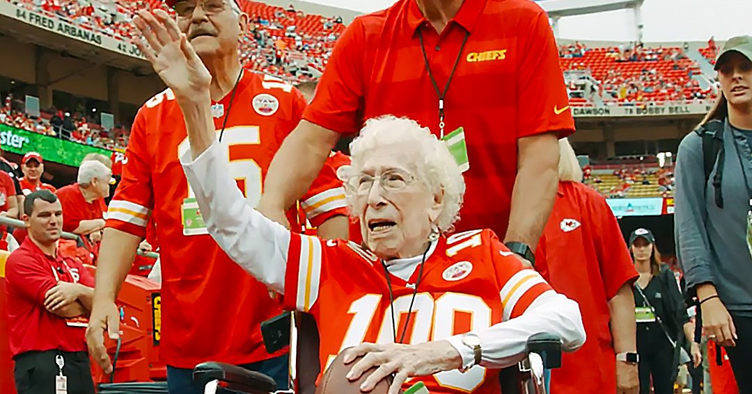 An older adult woman in a stadium wearing a red Kansas City Chiefs jersey with the number 100, surrounded by other individuals also dressed for a football game.